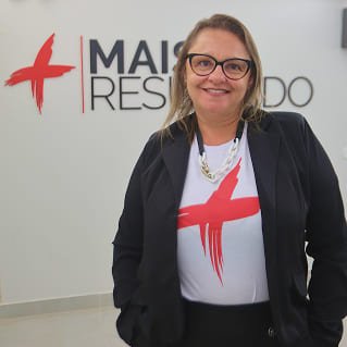 Alessandra Buzios, coordenadora de artes, sorrindo em frente ao logotipo da Agência Mais Resultado, vestindo camiseta personalizada e blazer preto.